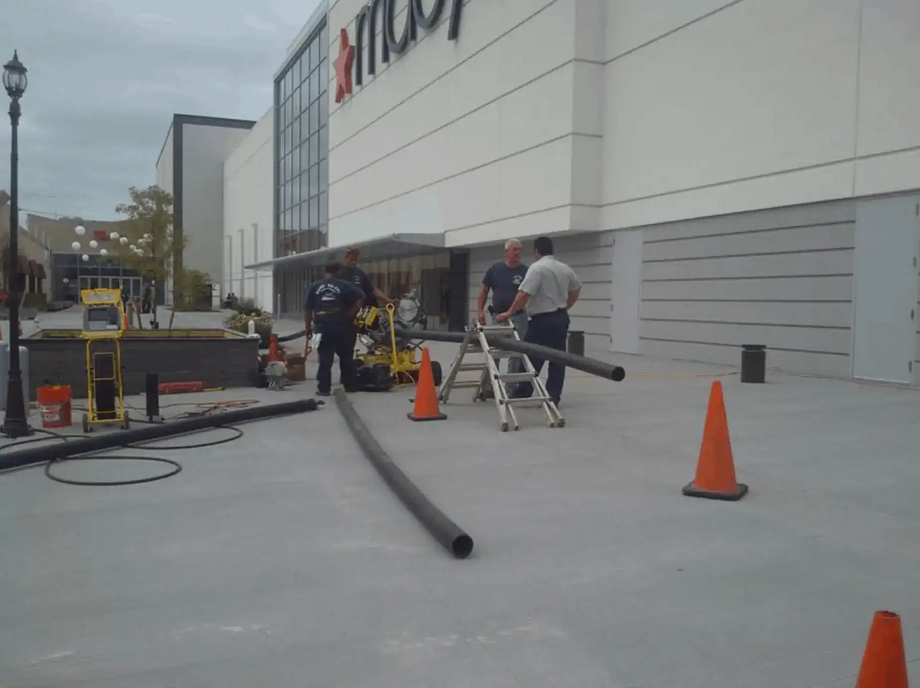 Construction work taking place outside a commercial storefront in Nassau County, NY, with scaffolding set up along the building’s facade, workers wearing safety vests and hard hats, construction materials and tools visible on the sidewalk, and a partially closed street with caution signs.