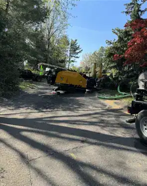 Roadwork machinery operating along a tree-lined street in Nassau County, New York, with construction equipment, traffic cones, and workers visible.