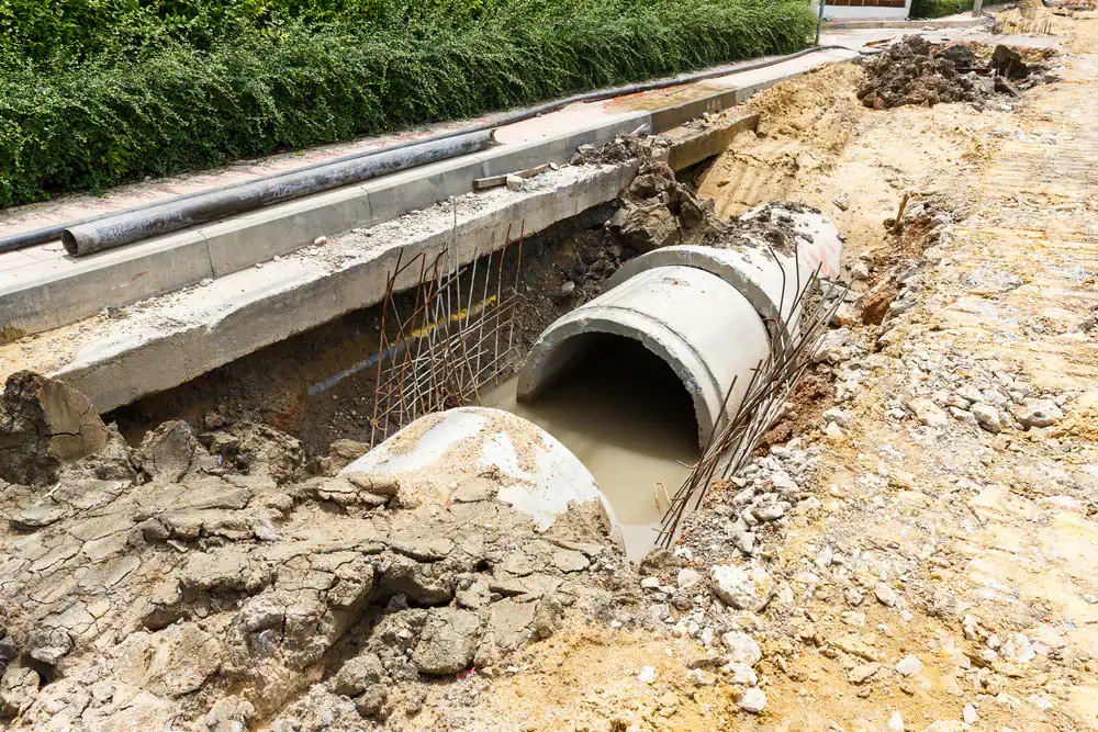 A construction site with large concrete drainage pipes partially installed in a trench filled with muddy water, surrounded by dirt, debris, and exposed rebar, next to a sidewalk lined with greenery.